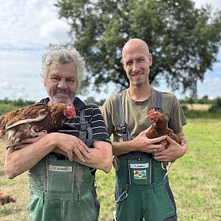Zwei Männer halten Hühner in einem Feld.